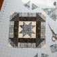 NYSNO New Snow White Snowflakes quilting cotton by Jason Yenter for In The Beginning Fabrics — featured as the background fabric in a vertical work-in-progress A Walk in the Woods quilt block on a white cutting mat in a bright sewing room, with dark brown mottled batik borders, blue-gray stripe star points, loose cut patches, a rotary cutter, and silver scissors. Quilt blocks shown for inspiration feature the A Walk in the Woods pattern — available at IraRobi Quilts.