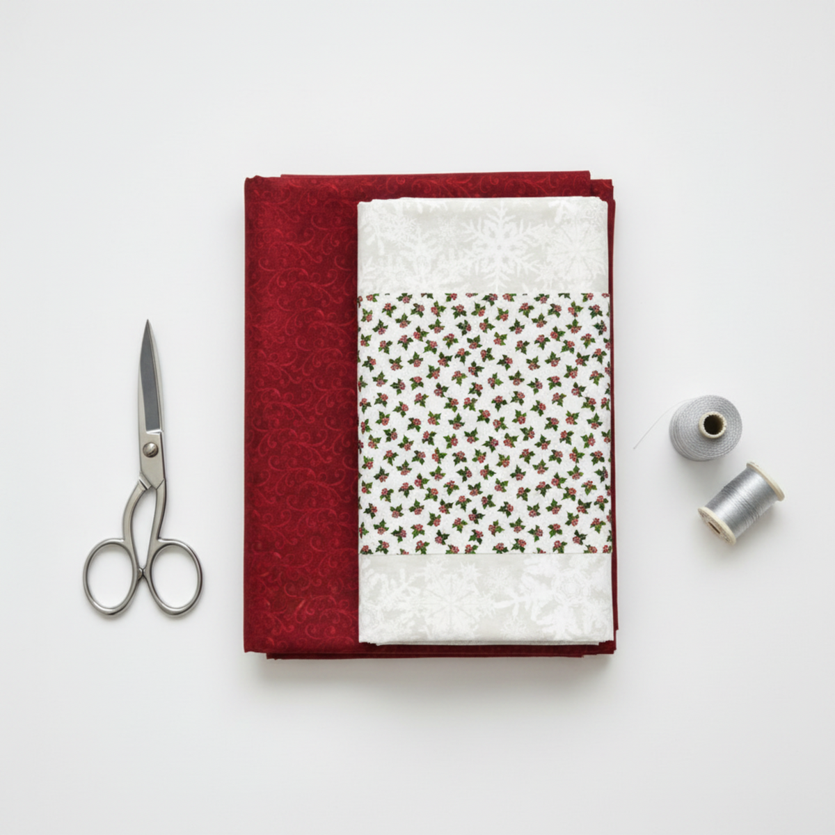 NYSNO New Snow Red Swirl quilting cotton by Jason Yenter for In The Beginning Fabrics — folded as the hero on a coordinating stack of winter holiday fabrics with silver scissors and silver thread spools on a white surface. Coordinating fabrics and quilt patterns available at IraRobi Quilts.