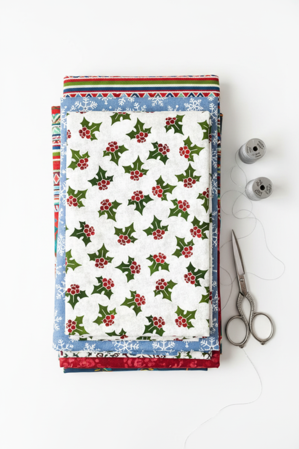 NYSNO New Snow White Holly quilting cotton by Jason Yenter for In The Beginning Fabrics — vertical flat lay folded as the hero on a tall stack of coordinating winter fabrics with silver thread spools and silver scissors on a white surface. Coordinating fabrics and quilt patterns available at IraRobi Quilts.