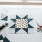 NYSNO New Snow White Holly quilting cotton by Jason Yenter for In The Beginning Fabrics — featured as the center fabric in a vertical work-in-progress A Walk in the Woods quilt block on a white cutting mat in a bright sewing room, with soft blue-gray mottled batik star points and background, loose cut patches, a rotary cutter, and silver scissors. Quilt blocks shown for inspiration feature the A Walk in the Woods Quilt Pattern by Beverly McCullough — available at IraRobi Quilts.