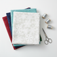 NYSNO New Snow White Snowflakes quilting cotton by Jason Yenter for In The Beginning Fabrics — folded as the hero on a stack of coordinating fabrics in deep red, navy, teal, and dark green, with silver thread spools and silver scissors on a white surface. Coordinating fabrics and quilt patterns available at IraRobi Quilts.