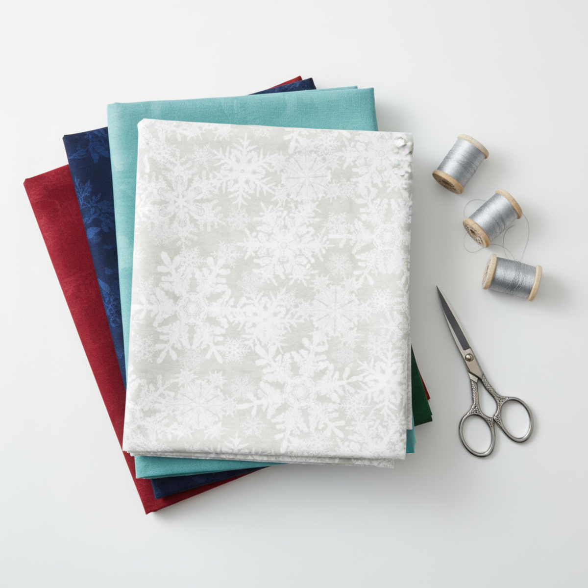 NYSNO New Snow White Snowflakes quilting cotton by Jason Yenter for In The Beginning Fabrics — folded as the hero on a stack of coordinating fabrics in deep red, navy, teal, and dark green, with silver thread spools and silver scissors on a white surface. Coordinating fabrics and quilt patterns available at IraRobi Quilts.