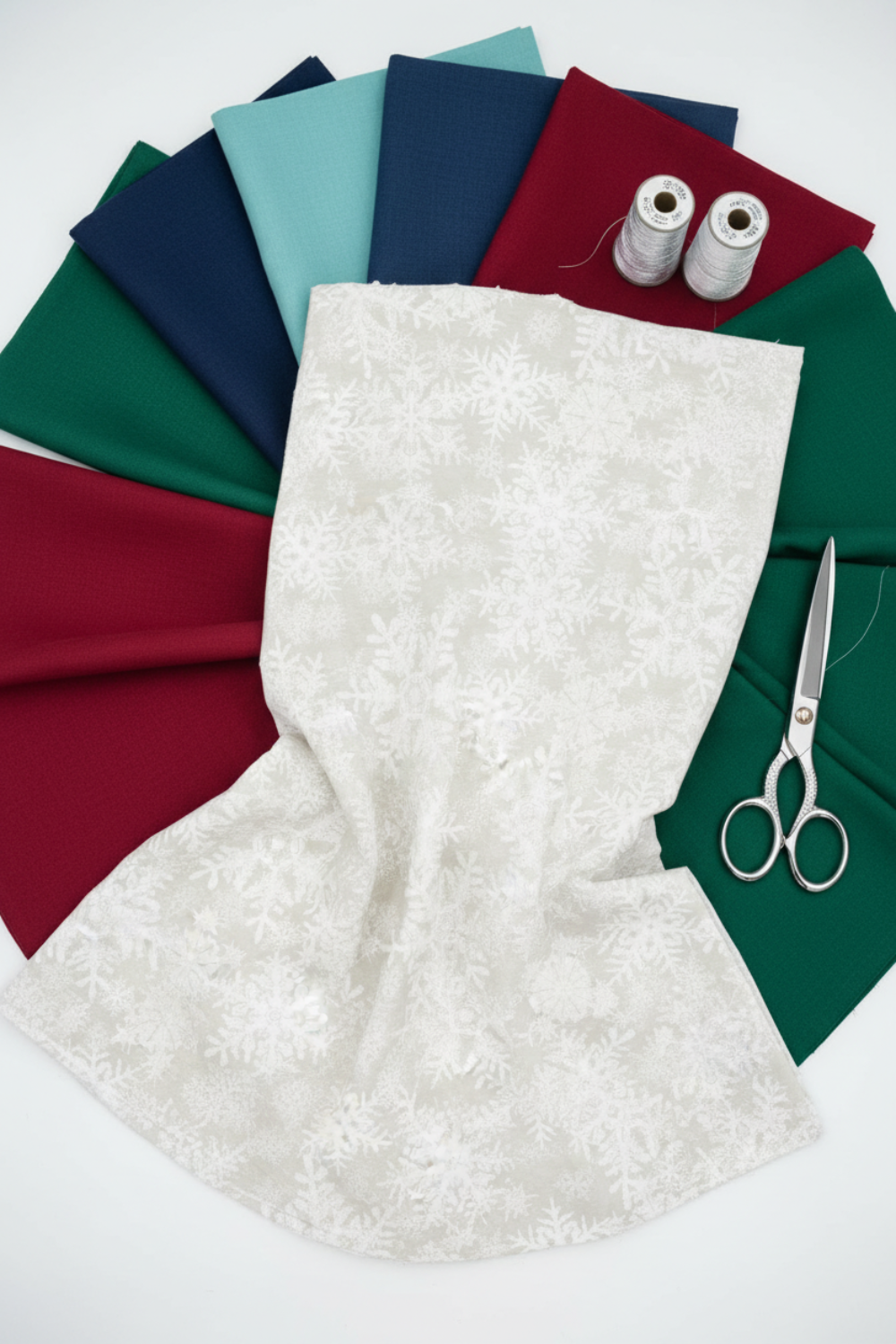NYSNO New Snow White Snowflakes quilting cotton by Jason Yenter for In The Beginning Fabrics — yardage draped as the front hero over a circular fan of coordinating fabrics in forest green, crimson, navy, teal, and dark green, with silver thread spools and silver scissors on a white surface. Coordinating fabrics and quilt patterns available at IraRobi Quilts.