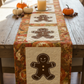 Oh Snap! gingerbread table runner by Jennie Jo Lamb for Lamb Farm Designs — three gingerbread person appliqué blocks on cream backgrounds with Autumn Celebration Multi Circles border fabric by Jason Yenter, featuring large medallion circles with autumn maple leaves in crimson, orange, and gold. Styled on a rustic farmhouse table with orange and white pumpkins, acorns, and a pillar candle. Quilts shown for inspiration feature the Oh Snap! pattern by Jennie Jo Lamb — available at IraRobi Quilts.