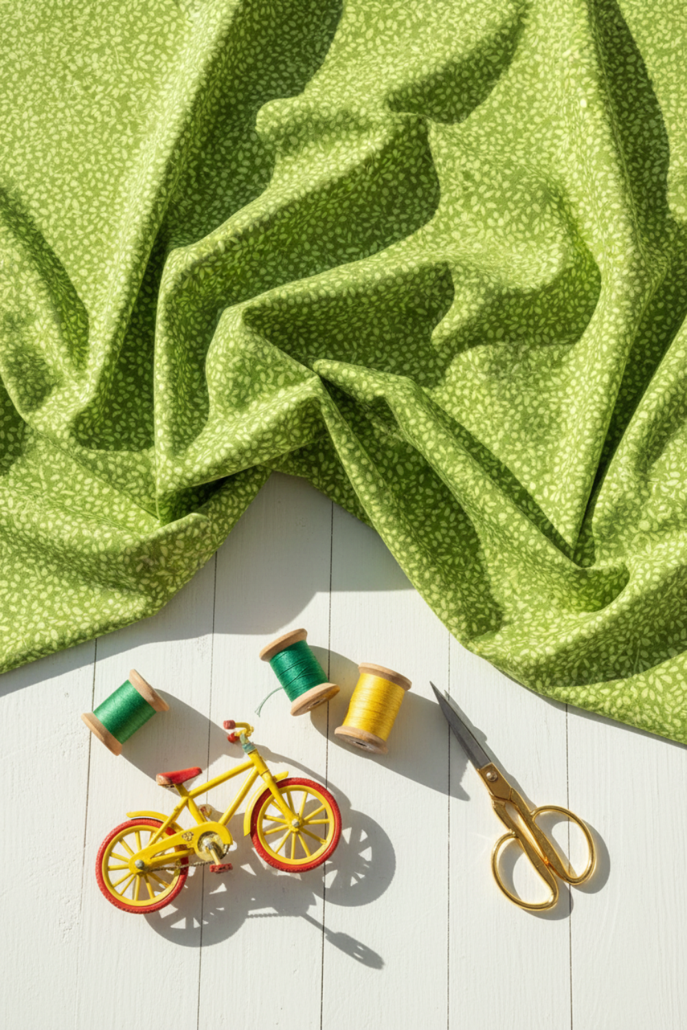 On the Go Leaves Green quilting cotton by Jason Yenter for In The Beginning Fabrics — vertical lifestyle photo with the tone-on-tone leaf print fabric draped as the hero on a white wood surface, with green and yellow thread spools, gold scissors, and a yellow toy bicycle below. Coordinating fabrics and quilt patterns available at IraRobi Quilts.
