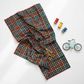 On the Go Plaid Multi quilting cotton by Jason Yenter for In The Beginning Fabrics — loosely draped and folded as the hero in a bright joyful quilting lifestyle flat lay with rainbow thread spools, gold scissors, and a small toy bicycle on a white surface.