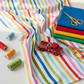 On the Go Stripes Multi quilting cotton by Jason Yenter for In The Beginning Fabrics — vertical lifestyle photo with the rainbow stripe as the clear hero in a joyful fan spread with rainbow thread spools and playful props. Coordinating fabrics and quilt patterns available at IraRobi Quilts.