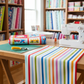 On the Go Stripes Multi quilting cotton by Jason Yenter for In The Beginning Fabrics — draped over a cutting table in a bright quilting studio with a white sewing machine, rotary cutter, and shelves of fabric bolts in the background. Coordinating fabrics and quilt patterns available at IraRobi Quilts.