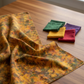 Expressions Batiks Hand-Dyes Orange Multi I fabric by Riley Blake Designs draped on a warm wood table with coordinating forest green, curry yellow, tomato red, and deep purple tonal batik fabric pieces nearby – complex multicolor hand-dyed batik with golden amber-orange base and olive green, slate blue-grey, and burnt red-orange, a rich moody focal blender perfect for nature-inspired and jewel-tone quilts, 100% cotton batik