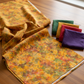 Expressions Batiks Hand-Dyes Orange Multi II fabric by Riley Blake Designs folded on a warm wood surface with coordinating forest green, tomato red, curry yellow, and deep purple tonal batik fabric pieces nearby – bright luminous golden amber-orange multicolor hand-dyed batik with red-orange splashes, olive green accents, and soft lavender-grey hints, a bold autumn harvest focal blender perfect for nature-inspired and jewel-tone quilts, 100% cotton batik, available at IraRobi Quilts
