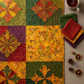 Autumn harvest quilt project flat lay featuring Expressions Batiks Hand-Dyes Orange Multi II fabric by Riley Blake Designs as the focal fabric – bright luminous golden amber-orange multicolor hand-dyed batik with red-orange splashes, olive green, and lavender-grey in pieced blocks with coordinating forest green, tomato red, curry yellow, and deep purple tonal batiks on a warm linen surface