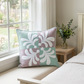 Pastel Harmony mint-and-mauve quilted pillow on a cream window bench beside an ivory bed and wooden nightstand with greenery.