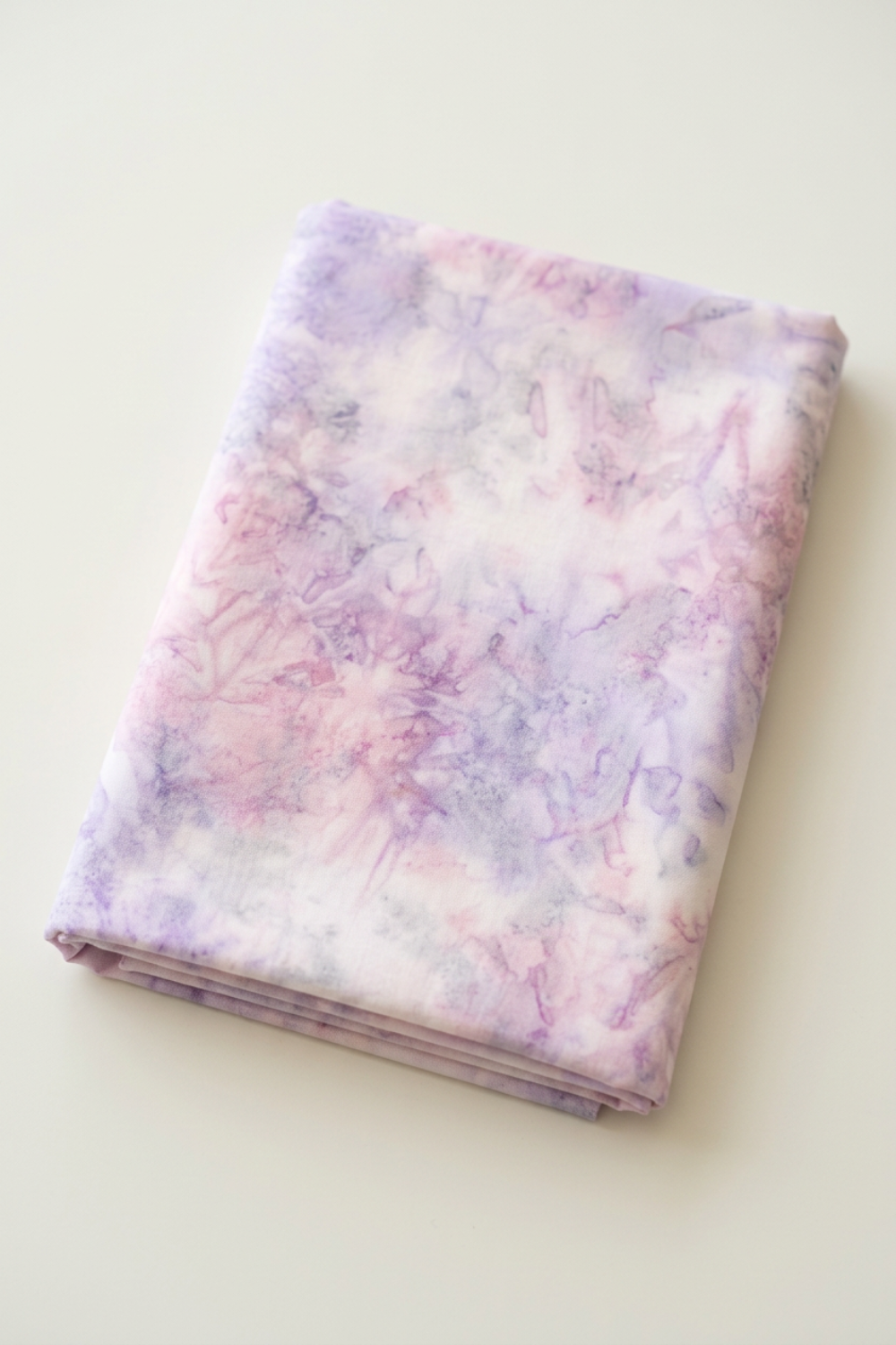 Neatly folded Color Play Mottled Lilac Lustre fabric by Riley Blake Designs on an ivory surface, showing pale pink and lavender mottling.