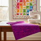 PAWsitivity Purple Field 3 Yard Cut quilting cotton by Jason Yenter for In The Beginning Fabrics — loosely draped in the foreground of a bright quilting studio with a colorful rainbow PAWsitivity half-square triangle quilt and Bernina sewing machine in the background. Quilts shown for inspiration feature the ModCat Quilt Pattern by Colourwerx — available at IraRobi Quilts.