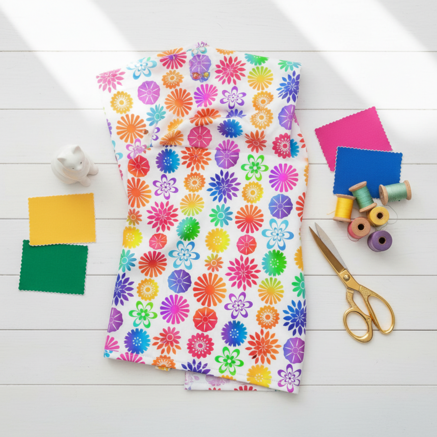 PAWsitivity White Flower Stripe quilting cotton by Jason Yenter for In The Beginning Fabrics — loosely draped on a white wooden surface with coordinating rainbow fabric swatches, rainbow thread spools, gold scissors, and a white ceramic cat figurine in a bright cheerful sewing room flat lay.