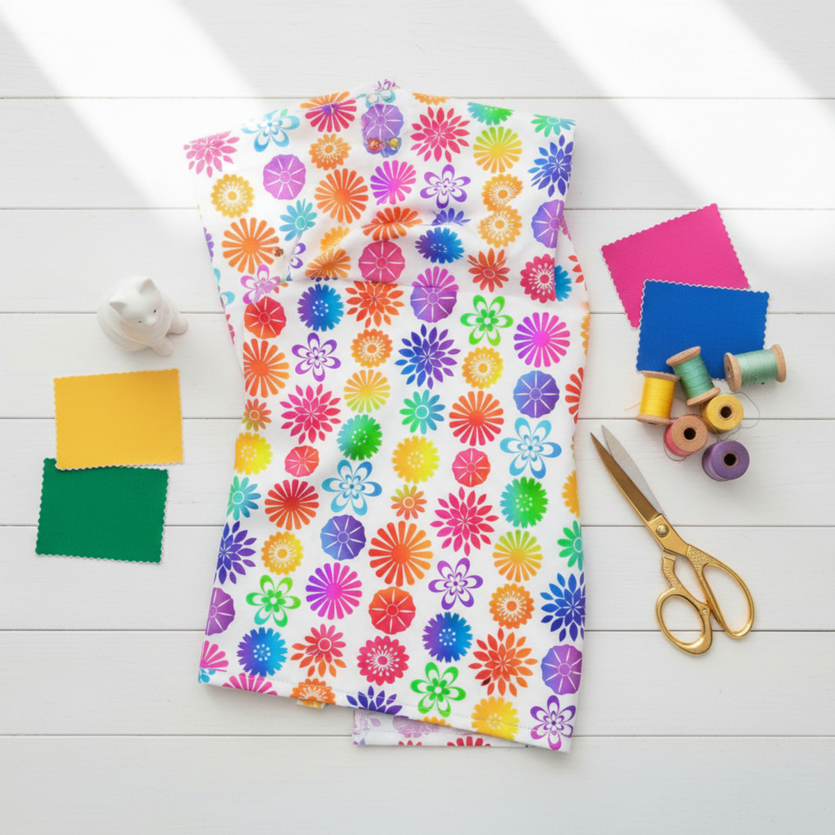PAWsitivity White Flower Stripe quilting cotton by Jason Yenter for In The Beginning Fabrics — loosely draped on a white wooden surface with coordinating rainbow fabric swatches, rainbow thread spools, gold scissors, and a white ceramic cat figurine in a bright cheerful sewing room flat lay.