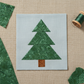 Pine tree appliqué quilt block featuring Expressions Batiks Hand-Dyes Evergreen fabric by Riley Blake Designs in three stacked triangle tiers – rich deep forest green hand-dyed batik with organic mottled crackle texture and sage green highlights on a soft blue-grey background, with warm brown batik trunk, loose cut Evergreen fabric pieces and green thread spool on a warm linen surface