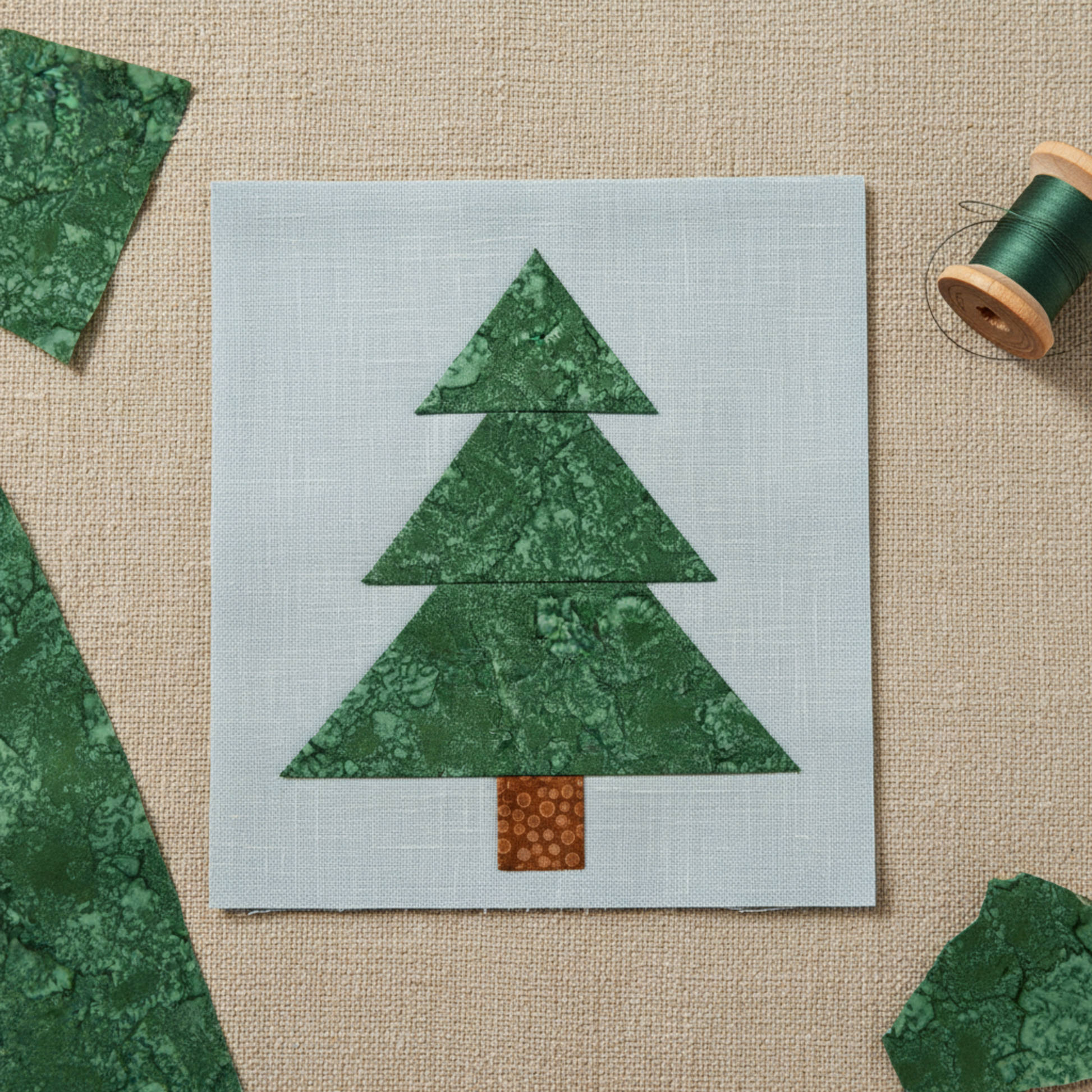 Pine tree appliqué quilt block featuring Expressions Batiks Hand-Dyes Evergreen fabric by Riley Blake Designs in three stacked triangle tiers – rich deep forest green hand-dyed batik with organic mottled crackle texture and sage green highlights on a soft blue-grey background, with warm brown batik trunk, loose cut Evergreen fabric pieces and green thread spool on a warm linen surface