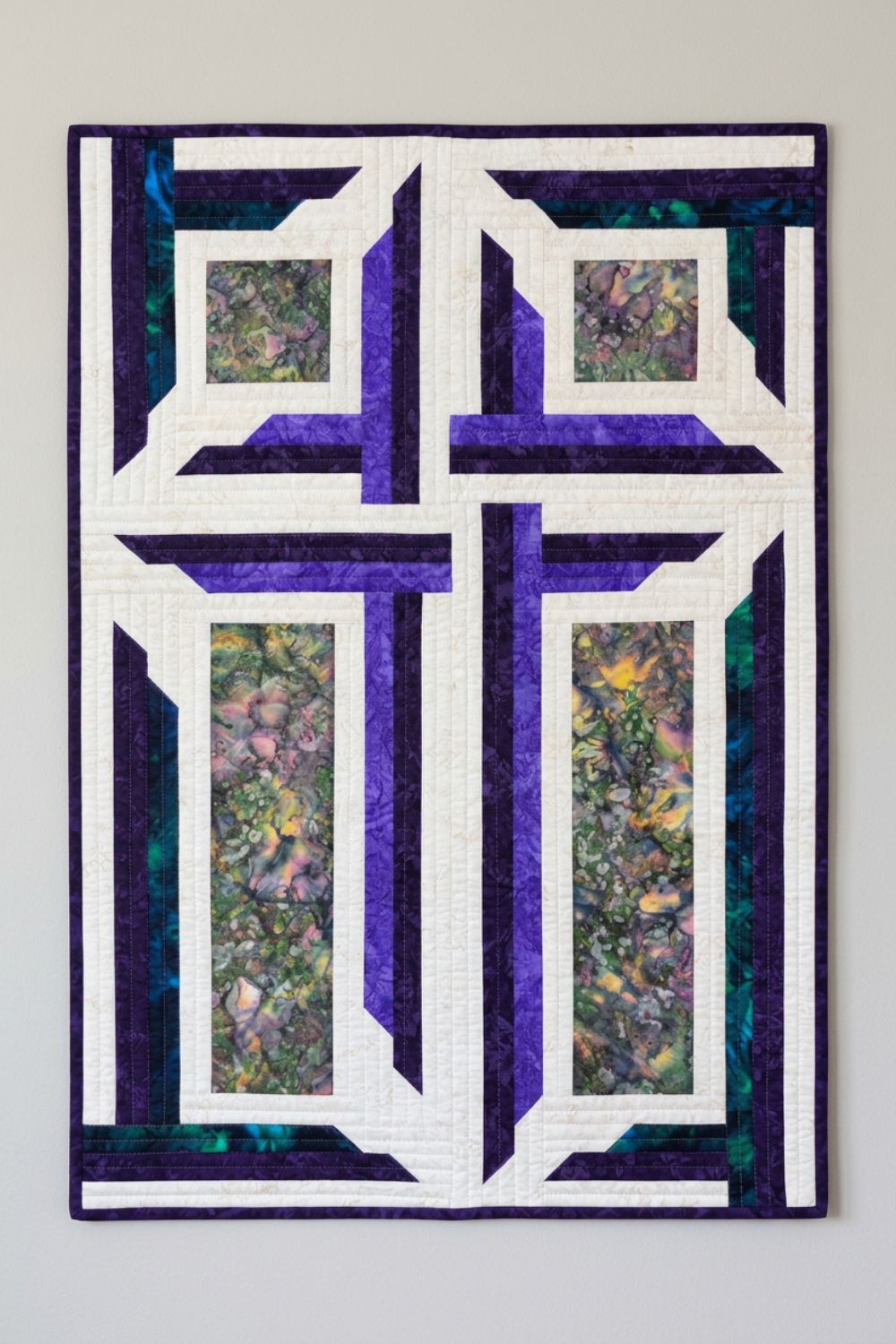 The Wondrous Cross handmade quilted wall hanging with a white cross, purple and teal patchwork, floral batik panels, and dark purple binding against a light-grey background.