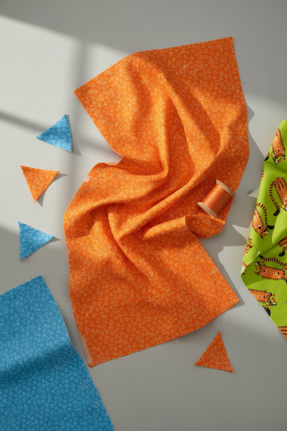 Safari Fun Floating Triangles Orange by Terry Runyan for Benartex – draped fabric with cut orange and blue triangle pieces, thread spool, and coordinating Tiger Too Lime fabric, natural window light