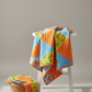 Safari Fun Floating Triangles Orange by Terry Runyan for Benartex – finished half-square triangle quilt in orange, blue, and green Safari Fun fabrics draped over a white stool with fabric scraps in a wicker basket