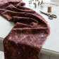 Poppy Seeds Raisin Fabric by Riley Blake Designs draped across a light-gray sewing table beside scissors, a thread spool, and dried flowers.
