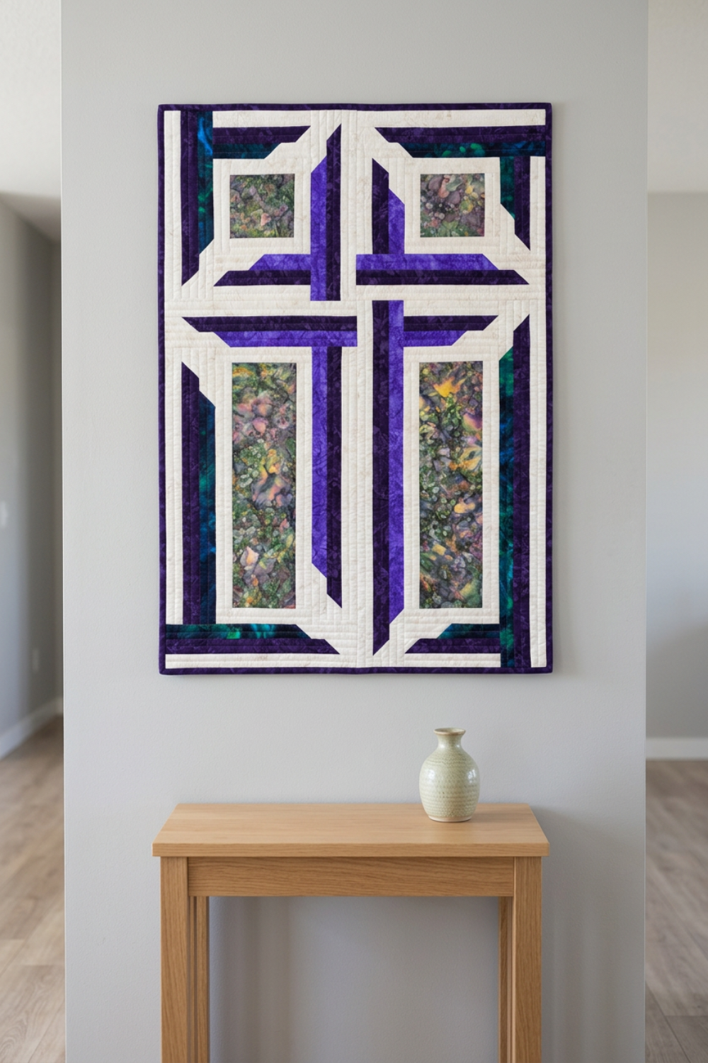 The Wondrous Cross purple, teal, and white cross wall quilt displayed above a small oak console with a pale ceramic vase in a light-grey entryway.