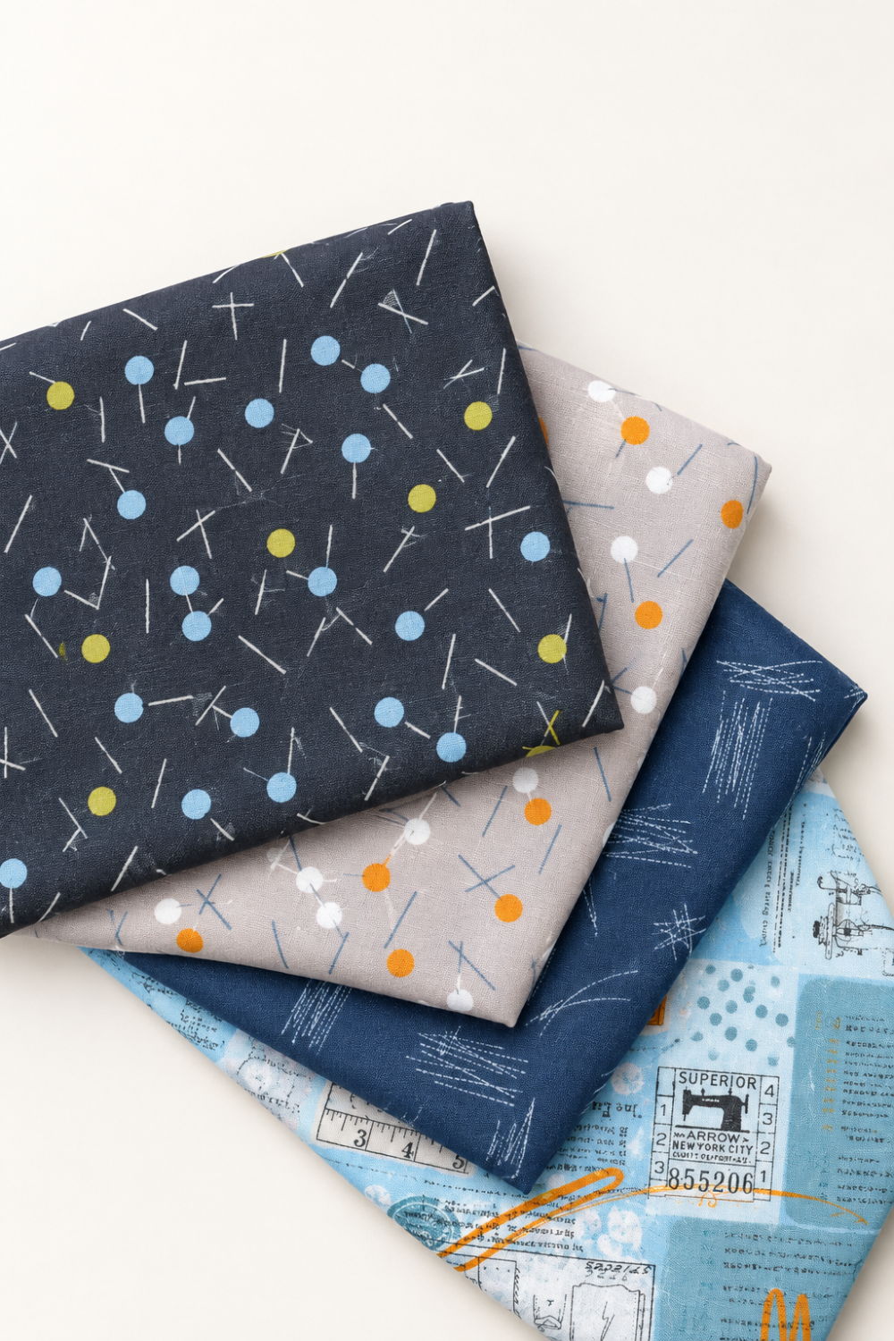 Vertical arrangement of folded coordinating fabrics in charcoal, gray, navy, and pale blue, topped with Bluish Pins & Buttons Charcoal.