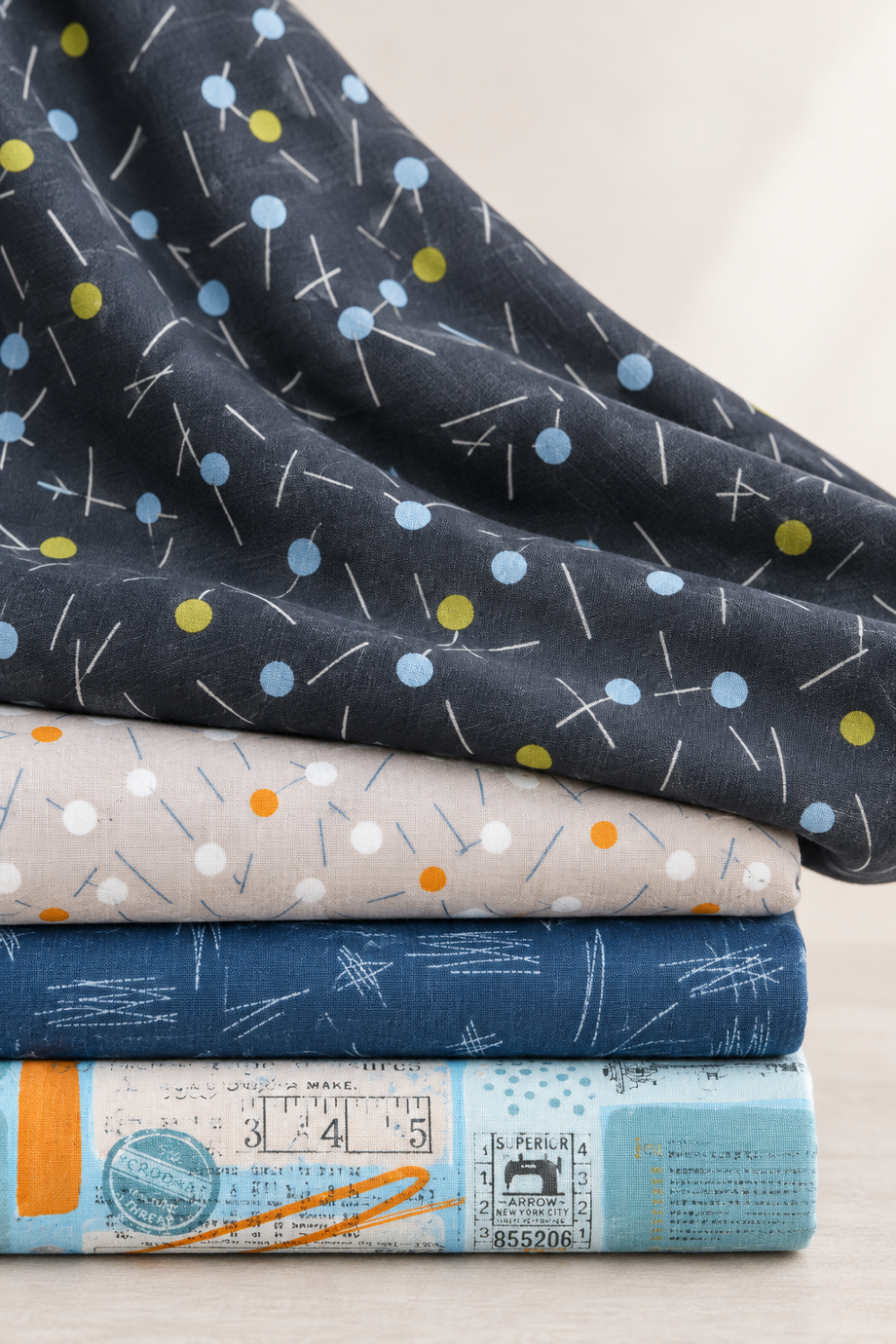 Bluish Pins & Buttons Charcoal fabric draped over a folded stack of coordinating gray, navy, and pale blue quilting fabrics.