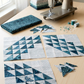 Three unfinished ocean-wave quilt blocks in assorted teal and pale fabrics on a sunlit sewing table with folded Terrazzo Smoky Teal batik, fabric scraps, pins, thread, and a sewing machine. Quilts shown for inspiration feature the Kelley Quilt Pattern by Edyta Sitar of Laundry Basket Quilts, available at IraRobi Quilts.