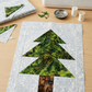 Unfinished green patchwork tree block with a Powdered Pearl print background lying flat on a wooden sewing table beside fabric remnants, pins, thread, and a sewing machine. Quilt blocks shown for inspiration. Coordinating fabrics and quilt patterns are available at IraRobi Quilts.
