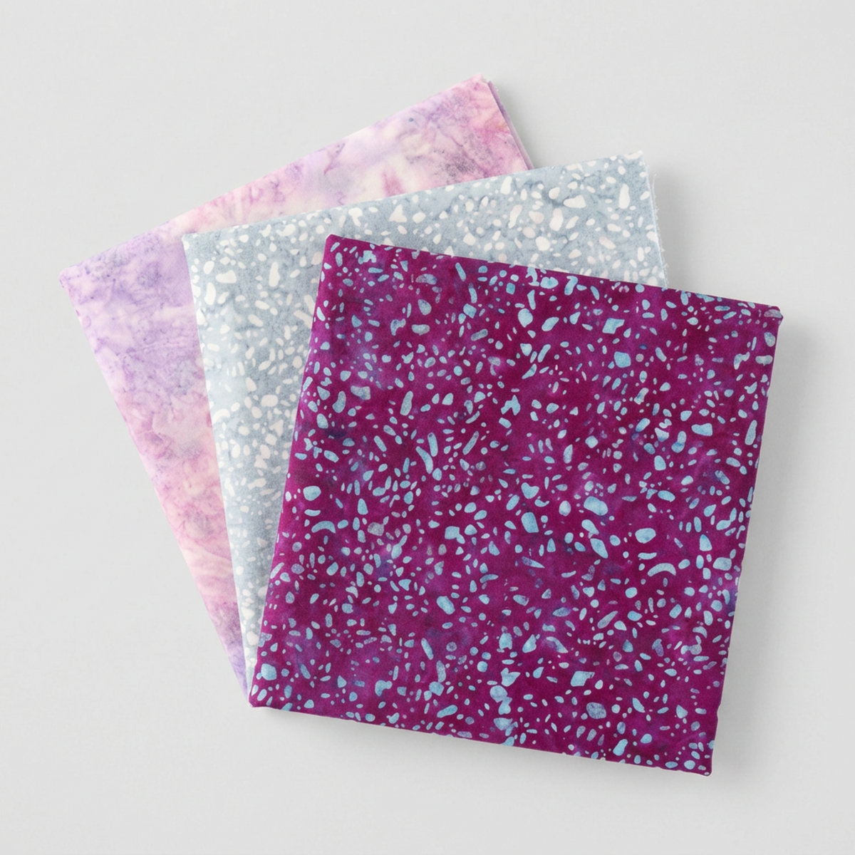 Terrazzo Purple Haze Fabric by Riley Blake Designs at the front of a three-fabric fan with coordinating pale blue-gray and pink-lilac fabrics.