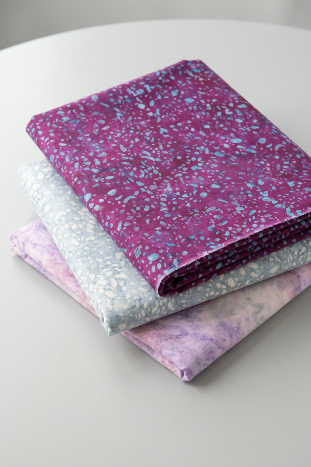 Terrazzo Purple Haze Fabric by Riley Blake Designs on top of a folded stack with coordinating pale blue-gray and pink-lilac fabrics.