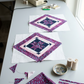 Two unfinished diamond-framed star blocks with Purple Haze accents lying on a sewing table beside fabric remnants, pins, thread, and a sewing machine. Quilt blocks shown for inspiration feature the Quartzite Quilt Pattern by Bear Hug Quiltworks, available at IraRobi Quilts.