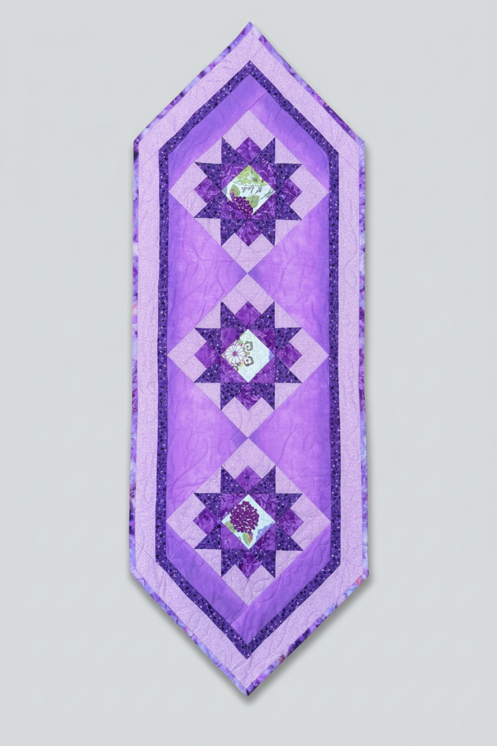 Bread and Butter Purple Twilight quilted table runner with three dark purple star blocks, floral centers, lavender borders and pointed ends on light gray.