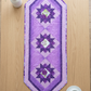 Purple Twilight quilted table runner centered lengthwise on a light wood tabletop beside a white vase of delicate stems and a white cup and saucer.