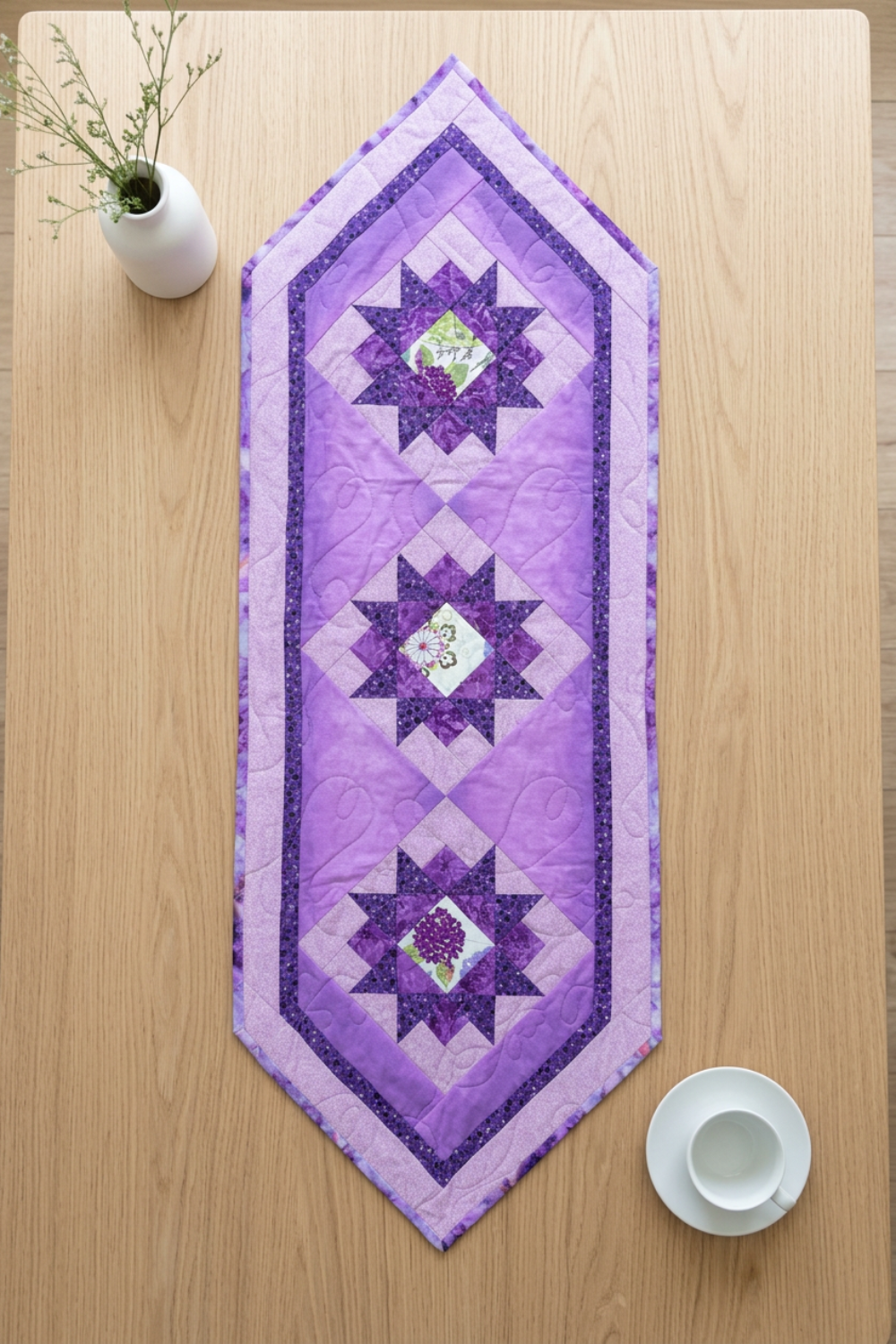 Purple Twilight quilted table runner centered lengthwise on a light wood tabletop beside a white vase of delicate stems and a white cup and saucer.