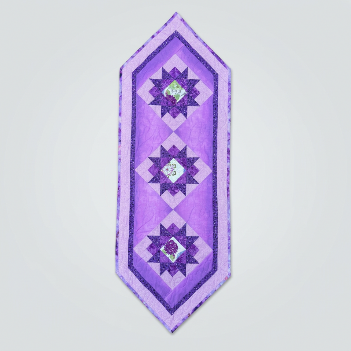Bread and Butter Purple Twilight quilted table runner with three dark purple star blocks, floral centers, lavender borders and pointed ends on light gray.