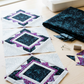 Three unfinished quilt blocks with Raven scrollwork centers, purple speckled accents, and pale surrounds on a sewing table beside folded Raven fabric, scraps, pins, thread, and a sewing machine. Quilts shown for inspiration feature the Quartzite Quilt Pattern by Bear Hug Quiltworks, available at IraRobi Quilts.