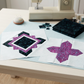 Unfinished quilt blocks in dark Raven scrollwork and purple speckled fabrics with light surrounds on a sewing table beside folded fabric, scraps, thread, and a sewing machine. Quilts shown for inspiration feature the Quartzite Quilt Pattern by Bear Hug Quiltworks, available at IraRobi Quilts.