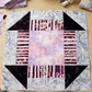 Quilt block with a Lilac Lustre center, plum striped bars, dark corner triangles, and gray botanical framing beside folded fabric, thread, pins, and a sewing machine. Quilt blocks shown for inspiration. Coordinating fabrics and quilt patterns are available at IraRobi Quilts.