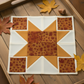 Autumn Celebration Brown Maple Leaves fabric by Jason Yenter for In The Beginning Fabrics featured as the center square and corner squares in a fall star quilt block — tossed dark burgundy maple leaf silhouettes on mottled amber and rust ground, with golden amber star points on cream background. Displayed on a rustic wood tray with scattered autumn maple leaves. Quilts shown for inspiration. Coordinating fabrics and quilt patterns available at IraRobi Quilts.