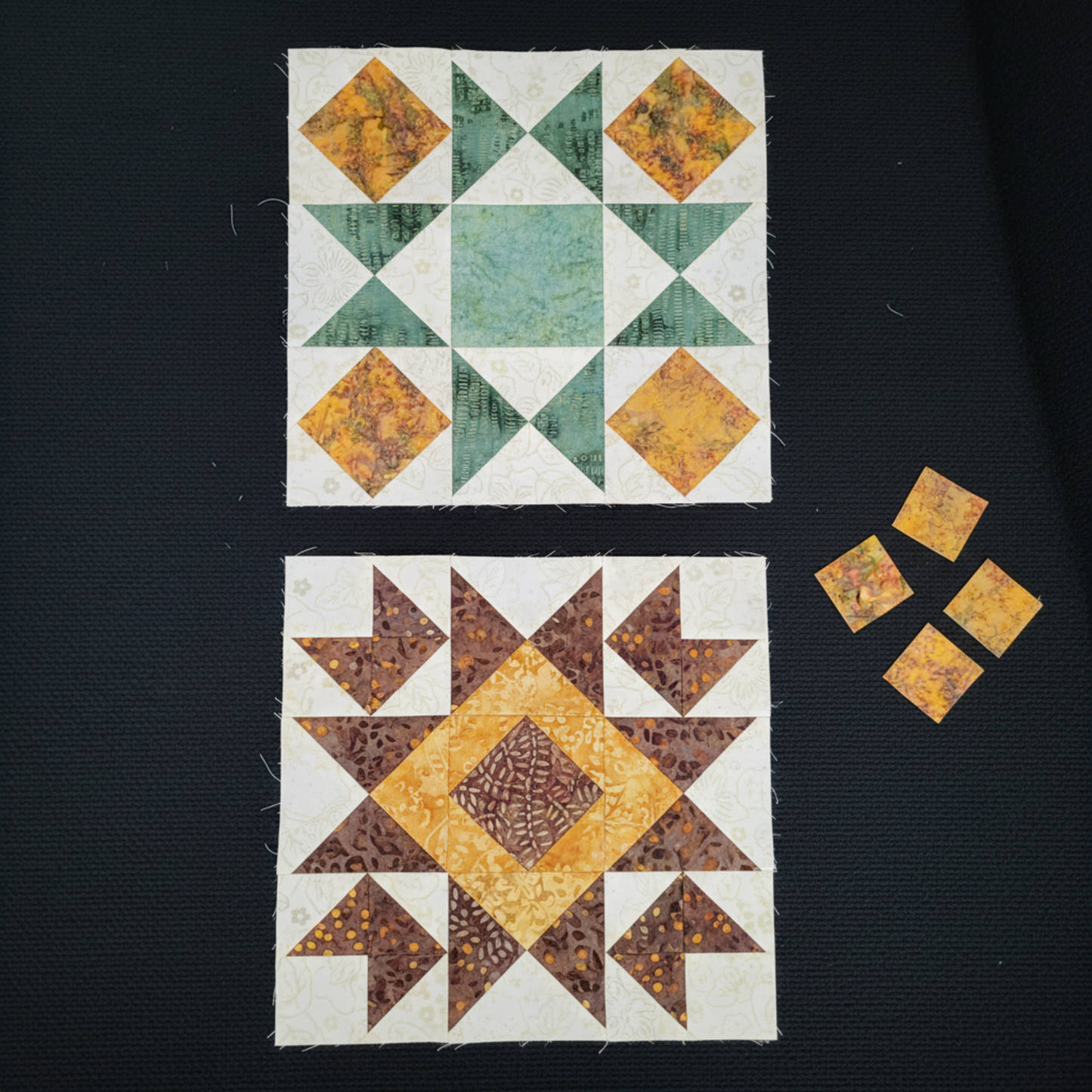 Two Hunter's Star quilt blocks work-in-progress on a dark black surface featuring Expressions Batiks Hand-Dyes Orange Multi I fabric by Riley Blake Designs – complex multicolor hand-dyed batik with warm golden amber-orange, olive green, slate blue-grey, and burnt red-orange in the star points and corner squares of both blocks, with teal-green and cream tonal batik in the top block and warm brown tonal batik in the bottom block, loose cut Orange Multi I fabric squares nearby