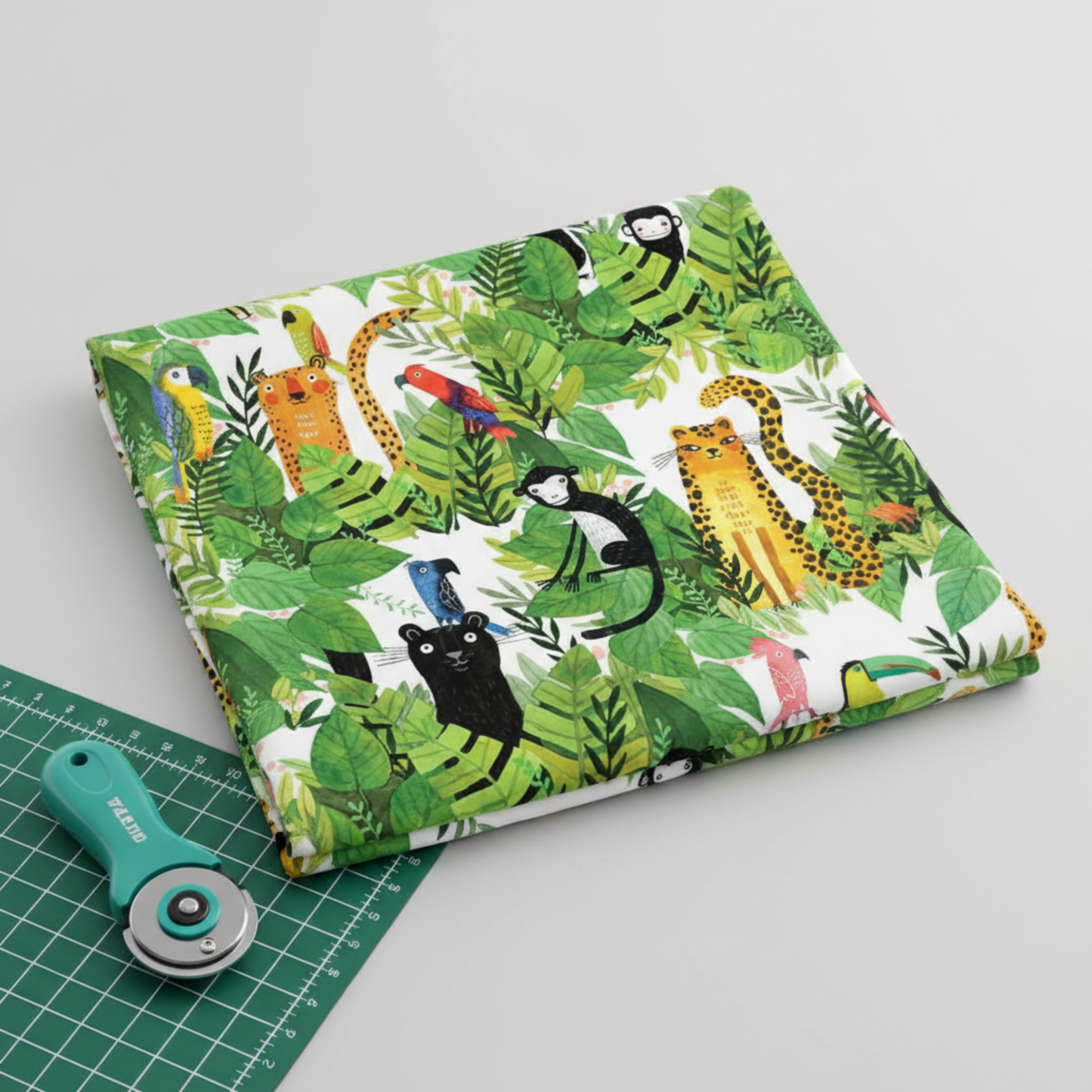 Safari Fun White/Multi quilting cotton partially folded on a light grey surface with a teal rotary cutter and corner of green cutting mat — large-scale jungle watercolor print by Terry Runyan for Benartex