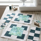 Two unfinished quilt blocks with Caribbean Shale daisy-print centers, light surrounds, and dark teal corner accents beside checkerboard sections, scraps, thread, and a sewing machine. Quilts shown for inspiration feature the Recipe Box Quilt Pattern by Bound to Be 