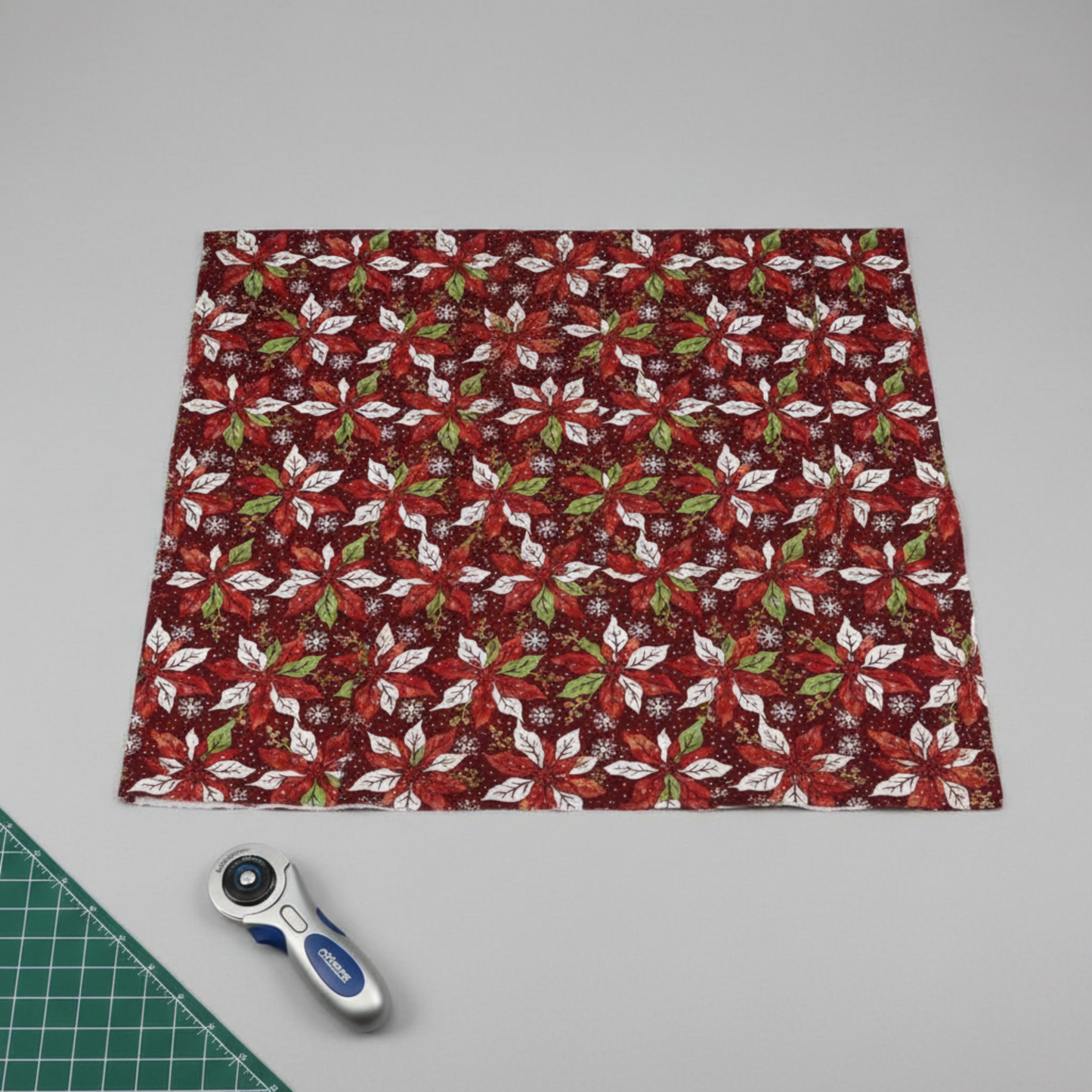 Winter Solstice II Red Poinsettia Metallic quilting cotton by Jason Yenter for In The Beginning Fabrics — fabric laid flat showing dramatic kaleidoscopic poinsettia print in deep red, white, and green on burgundy, shown in a clean modern quilting workspace with cutting mat and rotary cutter, 100% cotton with metallic accents. Quilt blocks shown for inspiration. Coordinating fabrics and quilt patterns are available at IraRobi Quilts.