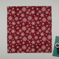 Winter Solstice II Red Snowflakes Metallic quilting cotton by Jason Yenter for In The Beginning Fabrics — fabric flat lay showing tossed white snowflake print on rich burgundy with gold metallic star dots, shown with cutting mat and rotary cutter in a clean quilting workspace, 100% cotton with metallic accents. Quilt blocks shown for inspiration. Coordinating fabrics and quilt patterns are available at IraRobi Quilts.