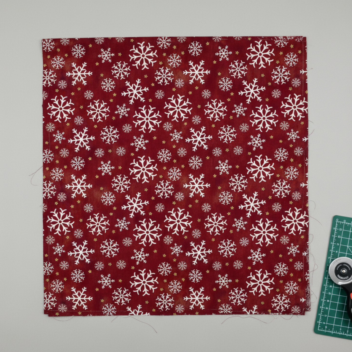 Winter Solstice II Red Snowflakes Metallic quilting cotton by Jason Yenter for In The Beginning Fabrics — fabric flat lay showing tossed white snowflake print on rich burgundy with gold metallic star dots, shown with cutting mat and rotary cutter in a clean quilting workspace, 100% cotton with metallic accents. Quilt blocks shown for inspiration. Coordinating fabrics and quilt patterns are available at IraRobi Quilts.