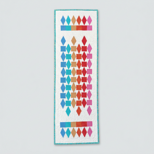 Refraction handmade quilted table runner with colorful diamonds and squares on a white background, turquoise binding and straight ends, displayed on light gray.