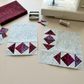 Two unfinished quilt blocks with burgundy and purple triangle-and-diamond units along the edges of large pale background areas, beside folded Mahogany fabric, pins, thread, and a sewing machine. Quilts shown for inspiration feature the Geese Over Galveston Bay Quilt Pattern by The Quilt Rambler, available at IraRobi Quilts.